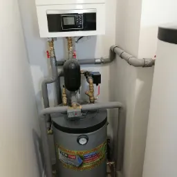 Kompaktowa kotłownia w pomieszczeniu: wiszący biały piec gazowy z czarnym panelem sterowania, szary zbiornik na wodę z etykietą producenta Eromet, rury z szarą izolacją, zawory i przyłącza.