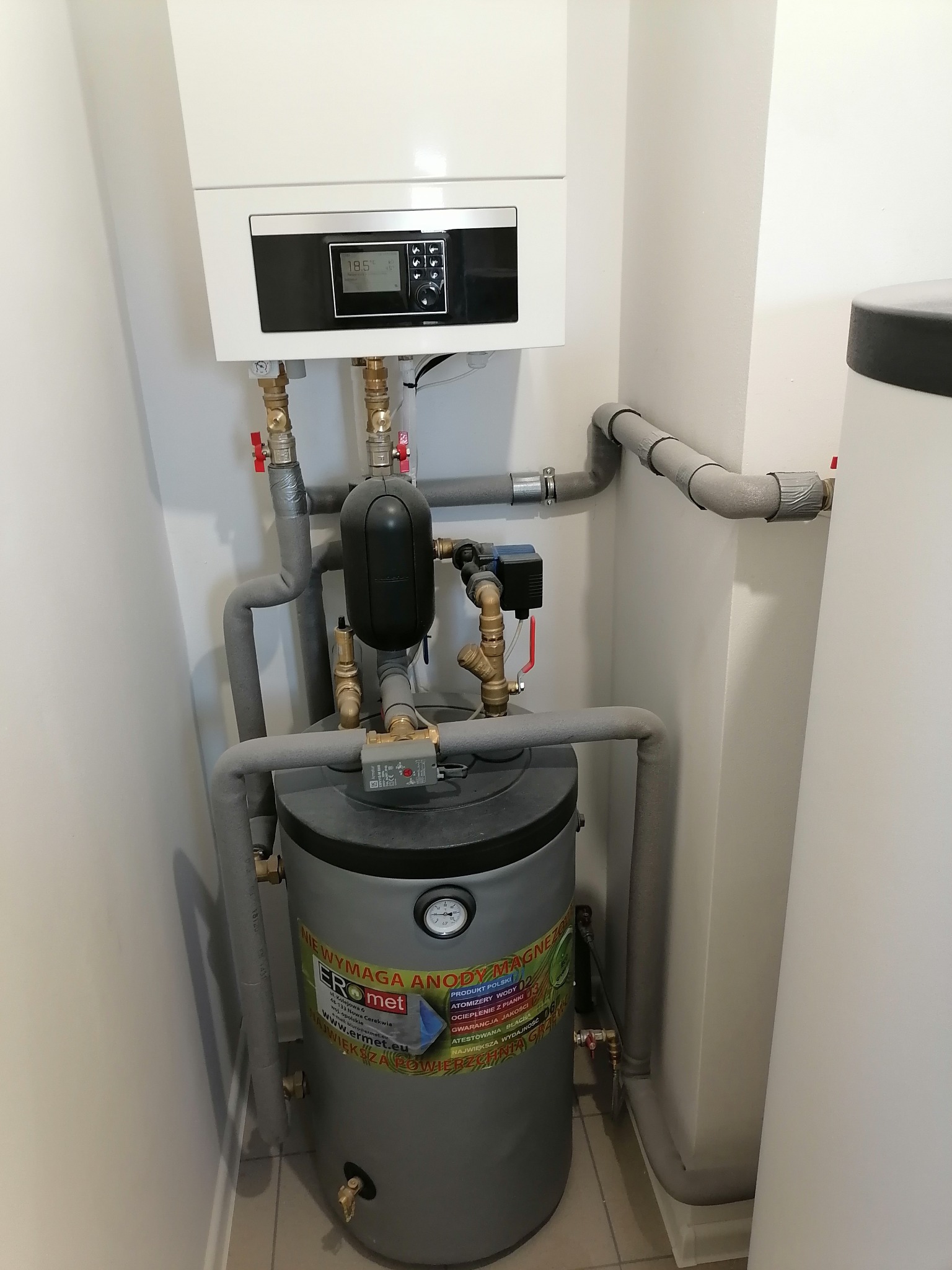 Kompaktowa kotłownia w pomieszczeniu: wiszący biały piec gazowy z czarnym panelem sterowania, szary zbiornik na wodę z etykietą producenta Eromet, rury z szarą izolacją, zawory i przyłącza.