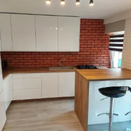 Nowoczesna kuchnia z białymi frontami szafek, drewnianym blatem i ścianą z czerwonej cegły, uzupełniona o wyspę z dwoma czarnymi hokerami.