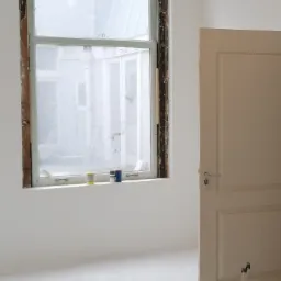 Pokój w trakcie odnawiania: okno z usuniętą ramą, trzy puszki farby na parapecie, białe ściany i drzwi z widocznymi kluczami.