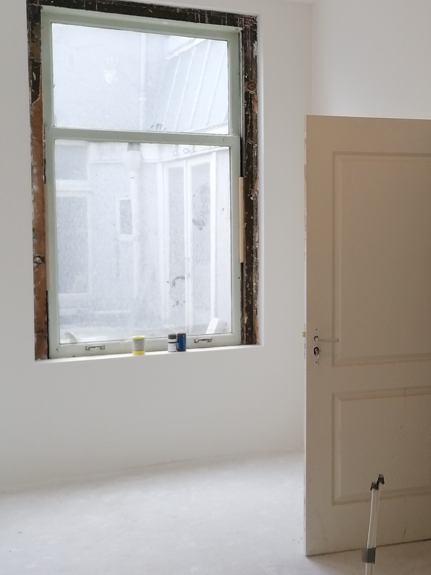Pokój w trakcie odnawiania: okno z usuniętą ramą, trzy puszki farby na parapecie, białe ściany i drzwi z widocznymi kluczami.