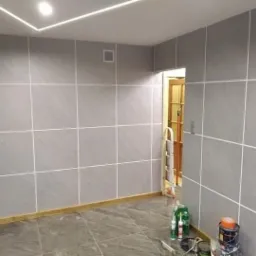 Wykończone wnętrze z szarymi płytkami na ścianach i podłodze, białe fugi, oświetlenie LED na suficie, widoczne narzędzia i materiały budowlane w rogu pomieszczenia.