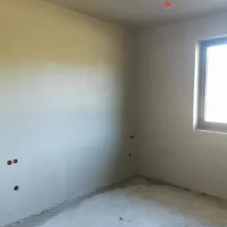 Pokój po nałożeniu gładzi gipsowej, widoczne puszki elektryczne i surowy beton na podłodze, okno z parapetem w prawym górnym rogu.