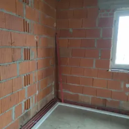 Wnętrze w trakcie budowy z widocznymi czerwonymi rurami instalacji elektrycznej ułożonymi wzdłuż ściany z cegły dziurawki, poziomica oparta o róg, surowy betonowy strop i fragment okna.