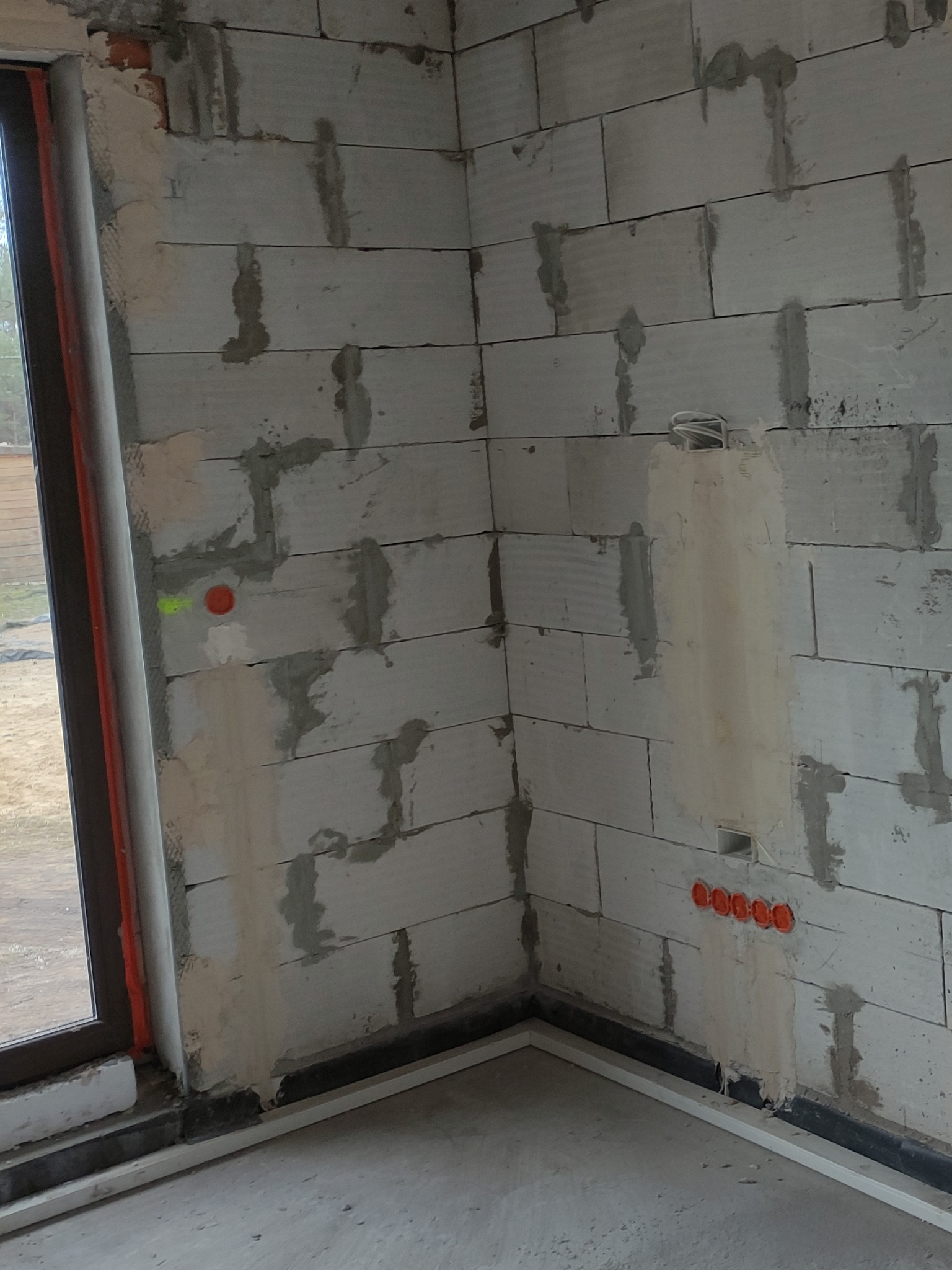 Surowy stan ściany z betonu komórkowego w rogu pomieszczenia z widocznymi puszkami elektrycznymi w kolorze pomarańczowym, przewodami i bruzdami wypełnionymi zaprawą.