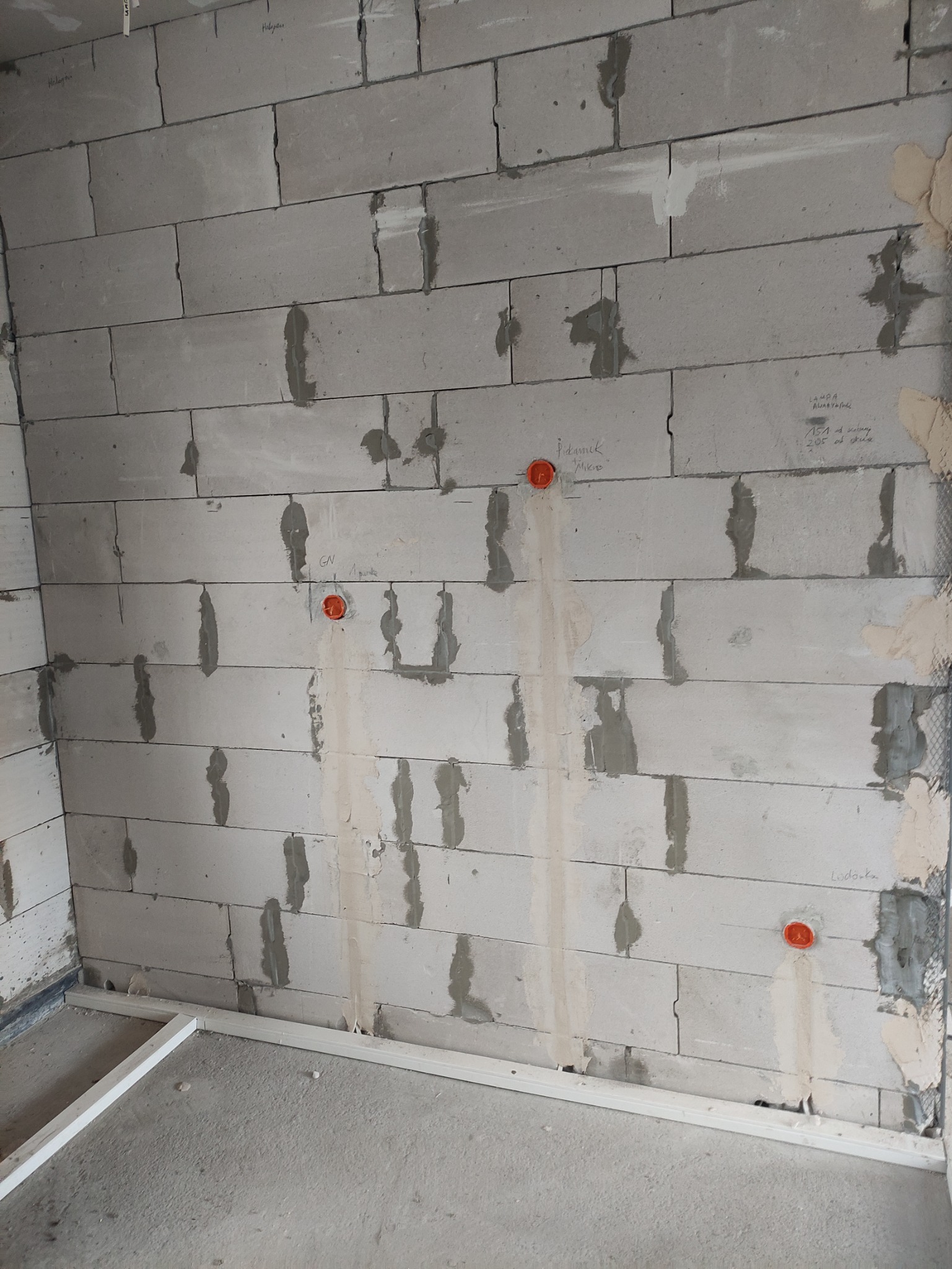 Ściana z betonu komórkowego w trakcie instalacji elektrycznej z widocznymi puszkami instalacyjnymi, częściowo wypełnionymi zaprawą, oraz odręcznymi notatkami na ścianie.