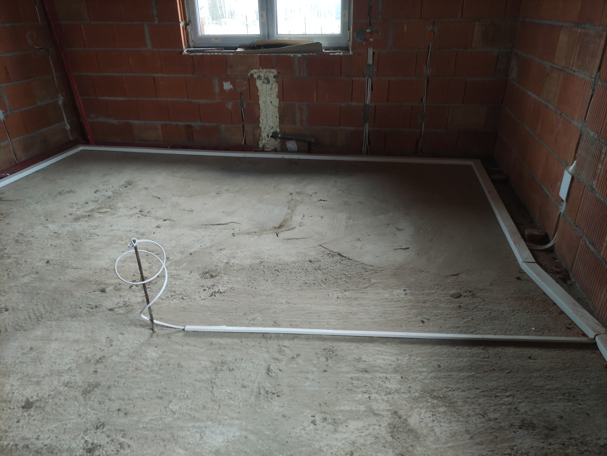 Surowy stan podłogi w nowym budynku z widocznymi przewodami elektrycznymi ułożonymi wzdłuż ścian i wystającym przewodem z podłoża, ceglane ściany i okno w tle.