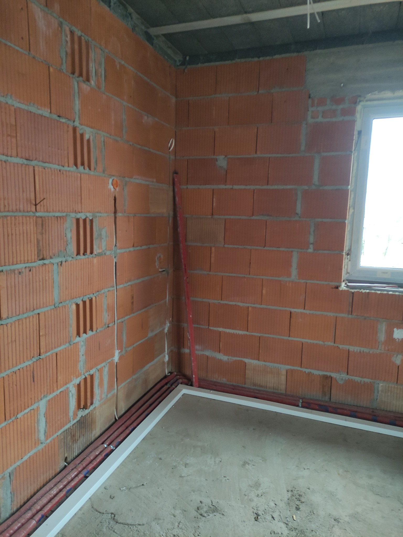 Wnętrze w trakcie budowy z widocznymi czerwonymi rurami instalacji elektrycznej ułożonymi wzdłuż ściany z cegły dziurawki, poziomica oparta o róg, surowy betonowy strop i fragment okna.