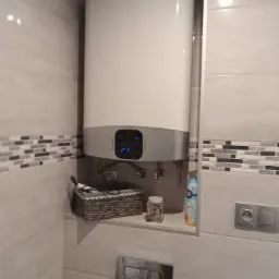 Bojler elektryczny Ariston zamontowany w łazience w Skwierzynie, widoczne przyłącza wodne, półka z koszykiem i ozdobami, płytki ceramiczne na ścianach z dekoracyjnym pasem mozaiki.