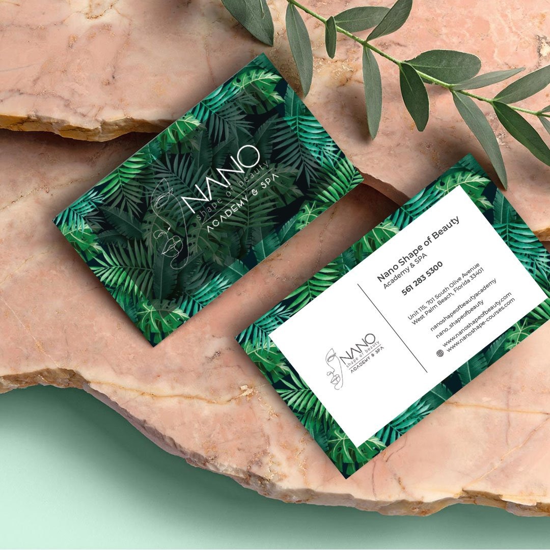 Dwie wizytówki Nano Shape of Beauty Academy & Spa z motywem tropikalnych liści, leżące na marmurowym blacie obok gałązki z zielonymi listkami.