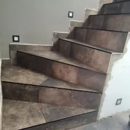 Schody wyłożone brązowo-szarymi płytkami ceramicznymi, z czarnymi krawędziami, widoczne małe, kwadratowe lampki w ścianie, ściana w odcieniu szarości, fuga w kolorze białym.