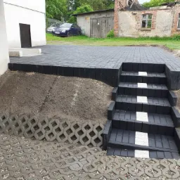 Ułożony z ciemnej kostki brukowej taras z czterostopniowymi schodami, wykończony jasnymi płytkami na każdym stopniu, oraz obrzeże z ażurowych płyt betonowych, na tle białego budynku i zarośniętego...