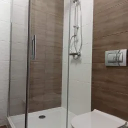 Kabina prysznicowa z chromowanymi elementami, biały brodzik, ściany wyłożone płytkami w odcieniach bieli i brązu, widoczna toaleta z przyciskiem spłukującym na ścianie.