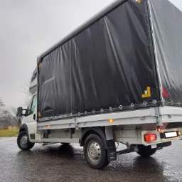 Biały samochód dostawczy z czarną plandeką na parkingu w pochmurny dzień, widok z tyłu i boku.