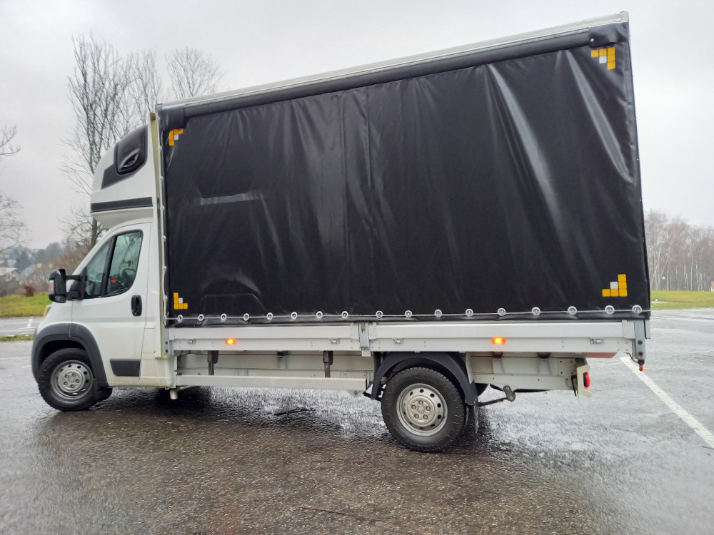 Biały samochód dostawczy z czarną plandeką na mokrym asfalcie, widok z boku.