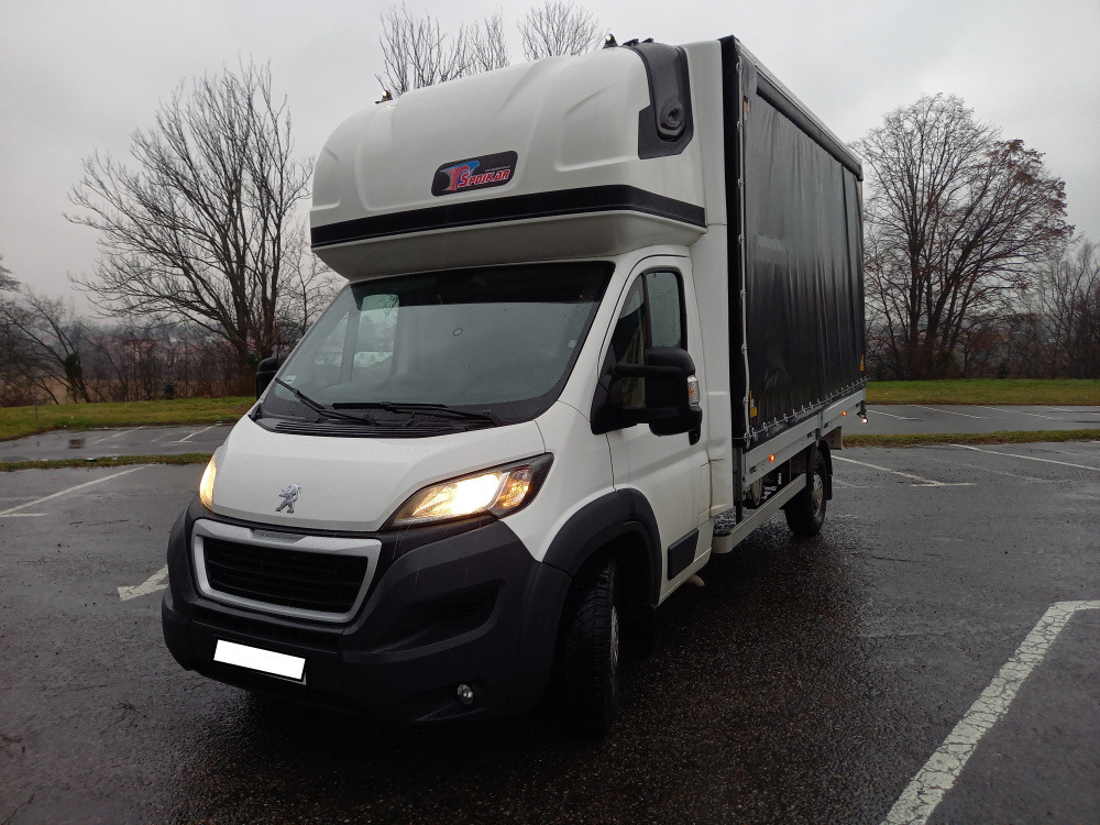 Biały samochód dostawczy Peugeot z plandeką na parkingu w pochmurny dzień, widoczne logo firmy Spokan na nadbudowie.