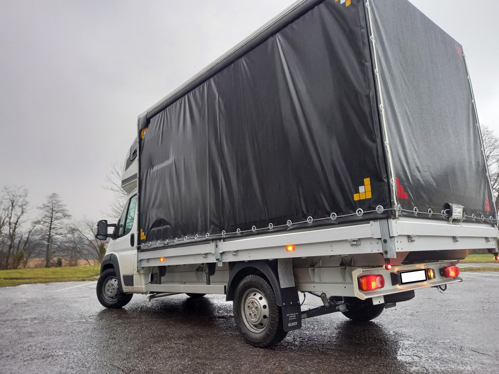 Biały samochód dostawczy z czarną plandeką na parkingu w pochmurny dzień, widok z tyłu i boku.