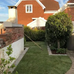 Świeżo założony trawnik z rolki w ogrodzie, wyrównywany grabiami, obrzeża wyłożone kostką, w tle dom z czerwonej cegły i zadbane krzewy.