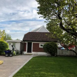 Pracownik w odzieży roboczej transportuje ziemię w wózku MAX po kostce brukowej na tle domu z czerwonej cegły i zielonego trawnika.