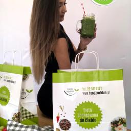 Catering Dietetyczny Blue - Kobieta z długimi włosami trzyma szklany słoik z zielonym koktajlem na tle toreb z logo firmy oferującej dietę pudełkową, widoczne warzywa i ściereczka w kratę.