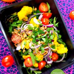 Catering Dietetyczny Blue - Sałatka z kalafiora, pomidorków cherry, czerwonej cebuli i orzechów włoskich, posypana świeżą natką pietruszki, w czarnym pojemniku na tle wzorzystej, różowo-niebieskiej serwety.