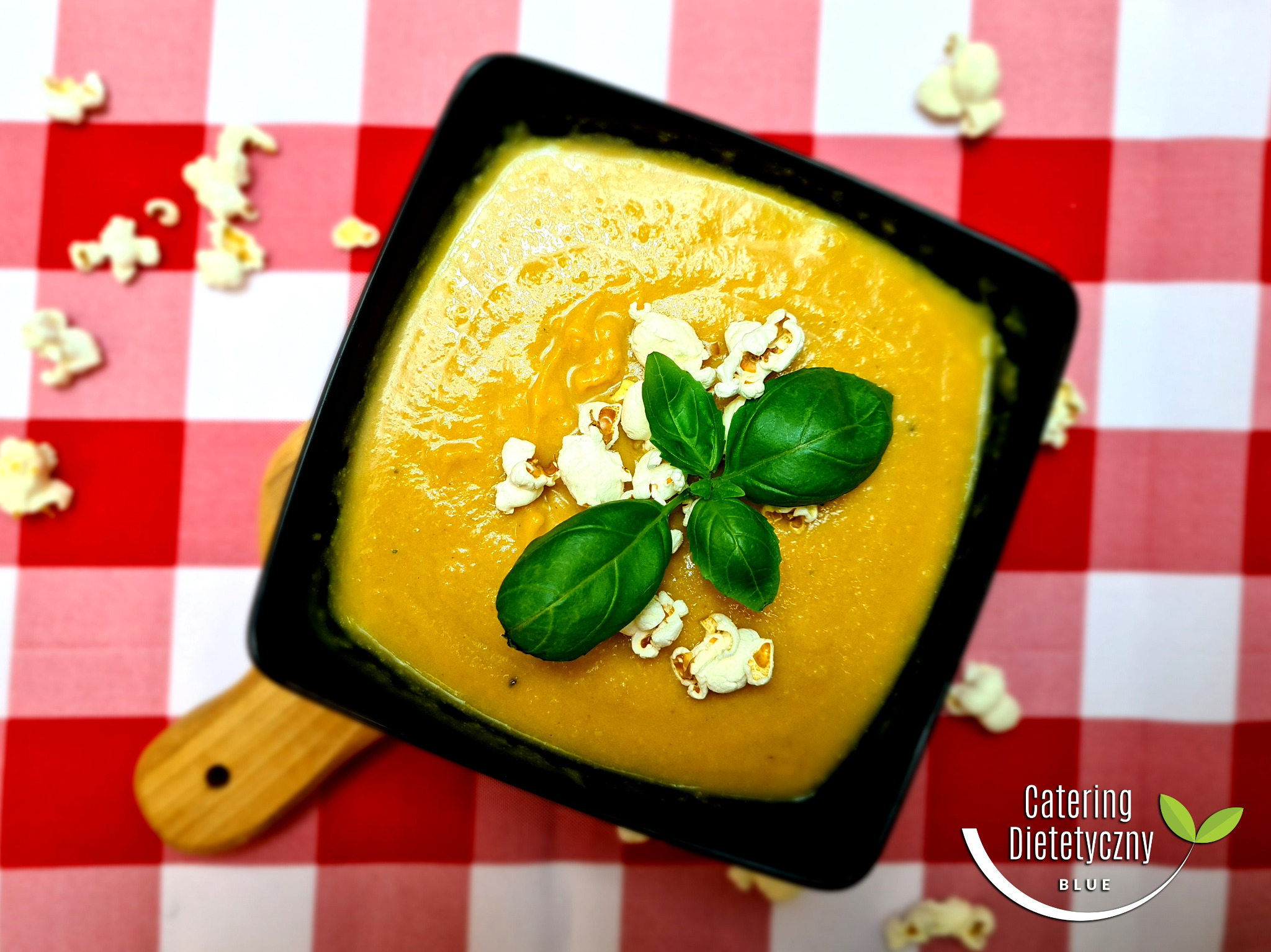 Krem z dyni w czarnym kwadratowym naczyniu, udekorowany świeżą bazylią i popcornem, na czerwono-białej kraciastej ściereczce.