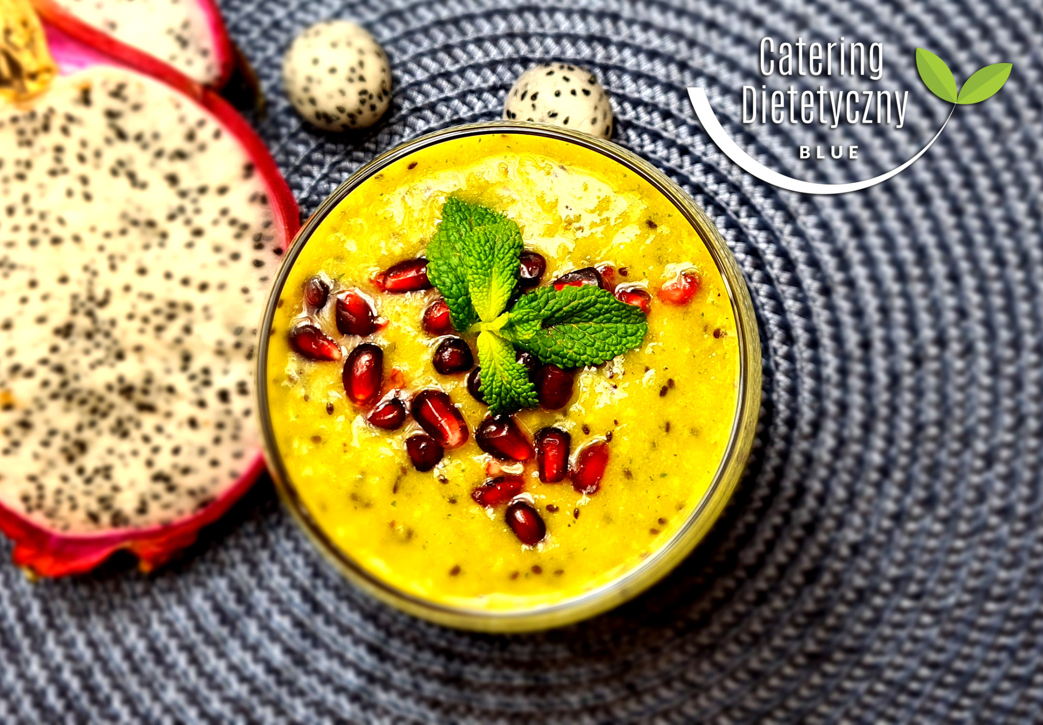 Żółty smoothie w szklance, udekorowany granatem i miętą, obok połówka pitai i dwa smocze owoce na szarym, plecionym tle. Widoczne logo firmy z nazwą.