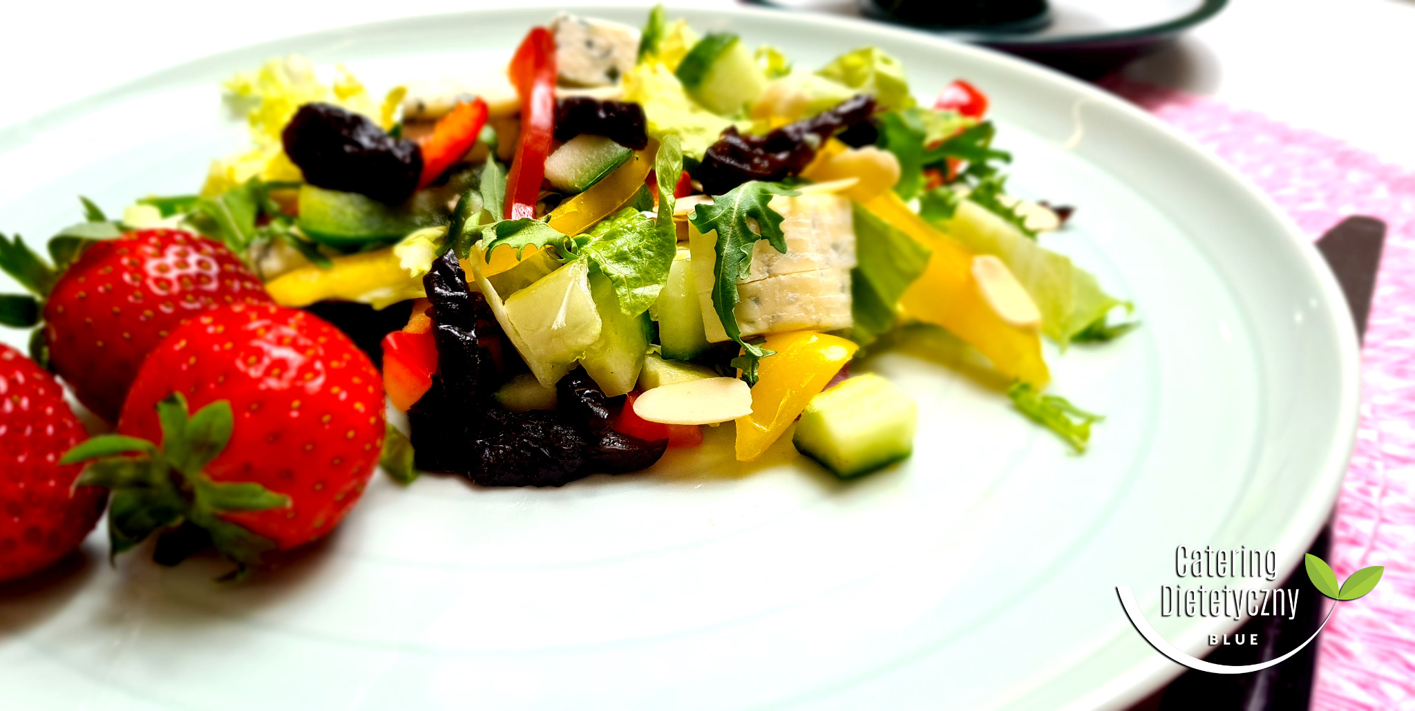Sałatka z truskawkami, serem pleśniowym, papryką i suszonymi śliwkami na białym talerzu, z logo firmy w prawym dolnym rogu.