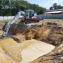 Raz Daw - Koparka Takeuchi na skraju wykopu wypełnionego wodą. Widoczne ślady pracy maszyny, ziemia i w tle budynek z białym ogrodzeniem. Drenaż terenu w trakcie realizacji.