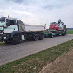 Raz Daw - Ciężarówka MAN z naczepą przewożąca czerwoną koparkę na zielonej platformie transportowej. Widok z boku na szosie, w tle pola i słupy wysokiego napięcia.