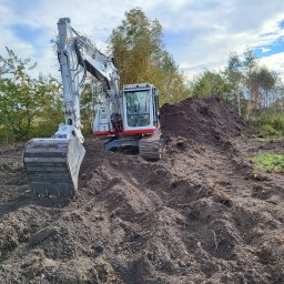 Raz Daw - Biało-szara koparka na nierównym, brązowym terenie z pryzmą ziemi w tle, pod błękitnym niebem z chmurami. Widoczne drzewa i fragment domu.