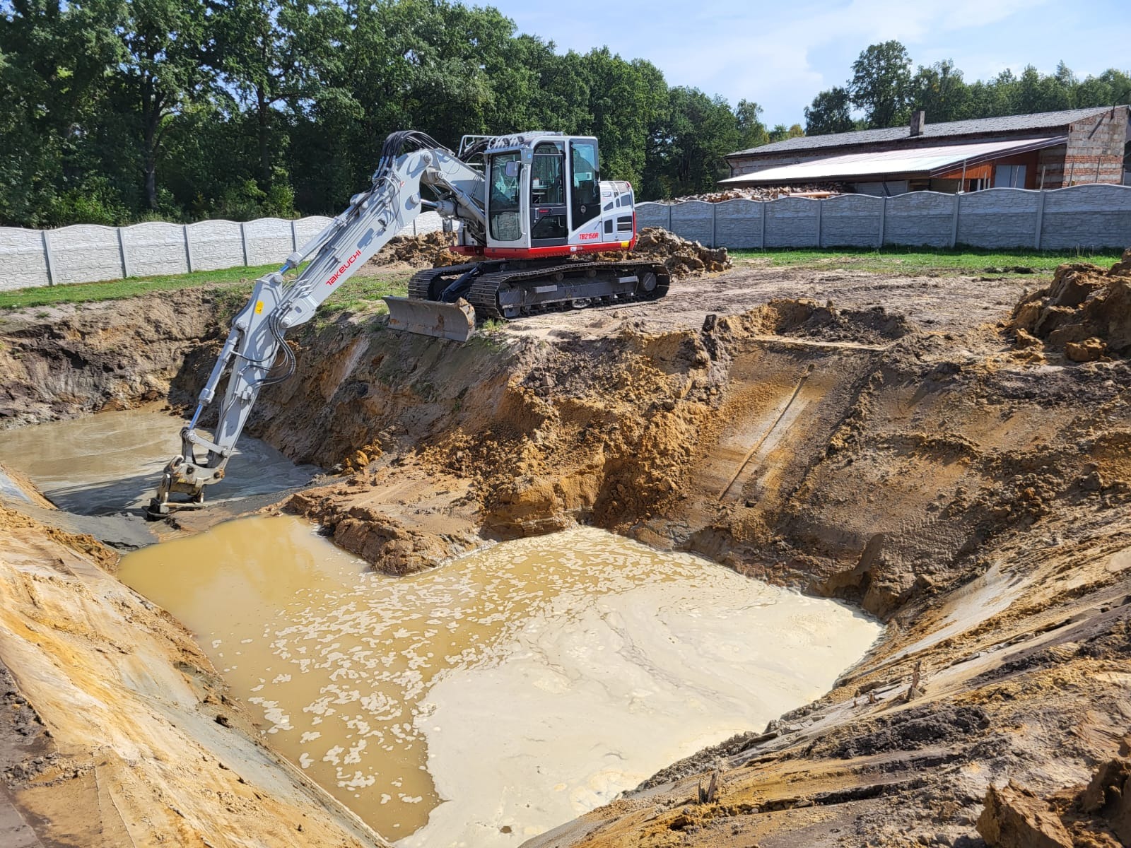 Koparka Takeuchi na skraju wykopu wypełnionego wodą. Widoczne ślady pracy maszyny, ziemia i w tle budynek z białym ogrodzeniem. Drenaż terenu w trakcie realizacji.
