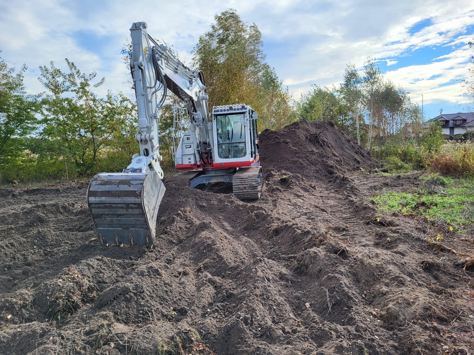 Biało-szara koparka na nierównym, brązowym terenie z pryzmą ziemi w tle, pod błękitnym niebem z chmurami. Widoczne drzewa i fragment domu.