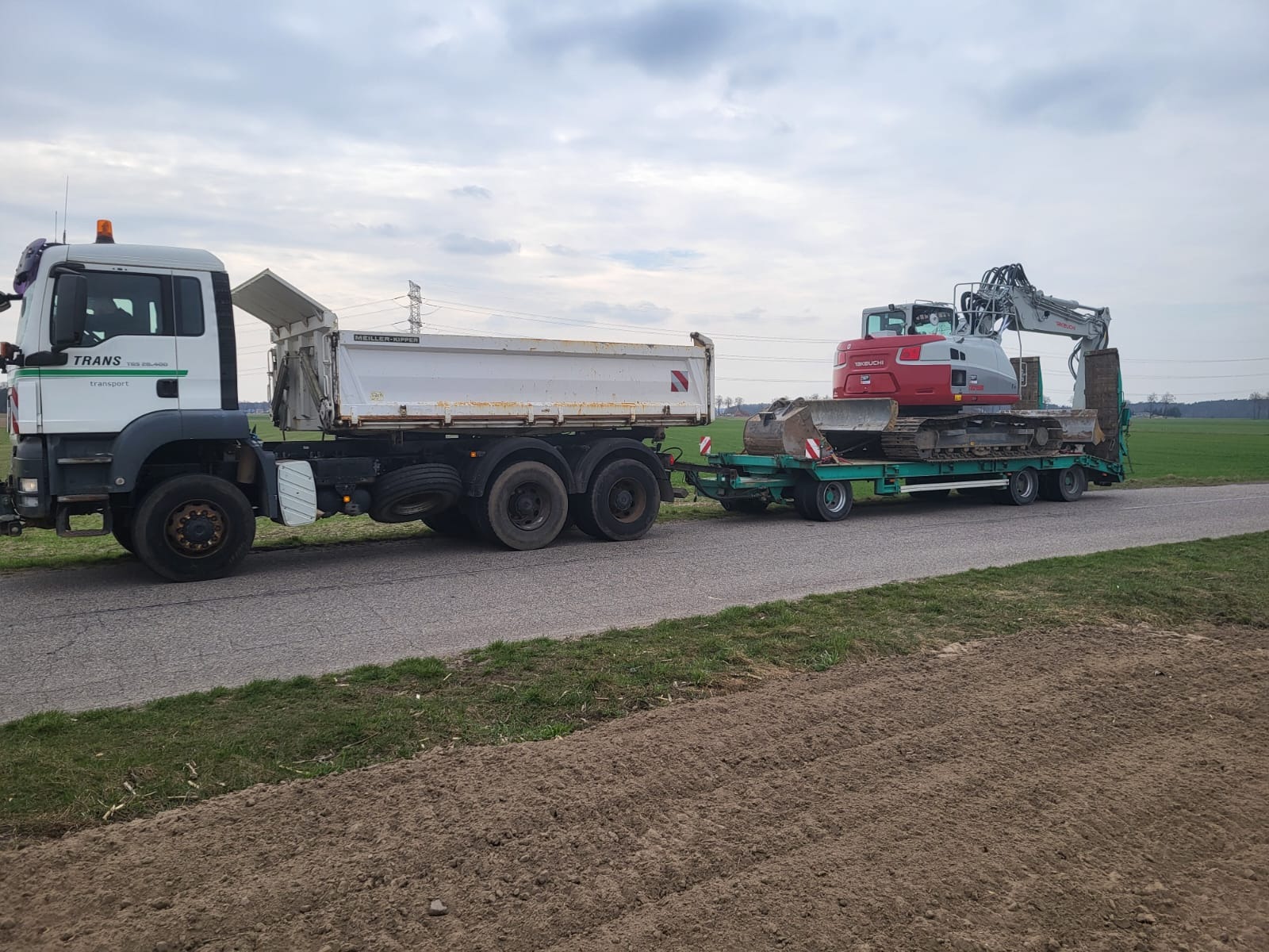 Ciężarówka transportuje koparkę Takeuchi na niskopodwoziowej przyczepie. Widok z boku na szosie, pole w tle. Pochmurne niebo.