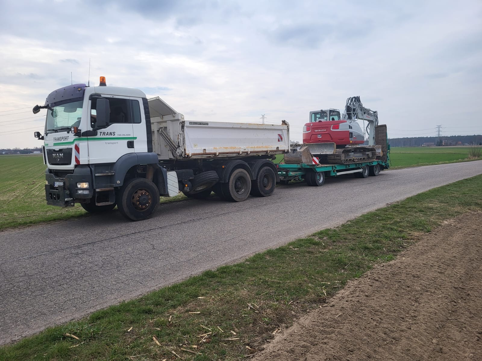Ciężarówka MAN z naczepą przewożąca czerwoną koparkę na zielonej platformie transportowej. Widok z boku na szosie, w tle pola i słupy wysokiego napięcia.