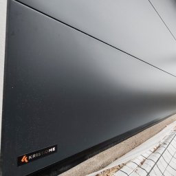 VINSTA SPÓŁKA CYWILNA WOJCIECH PAJĄK, DMYTRO ZHULINSKYI - Zamknięta, grafitowa brama garażowa segmentowa z widocznym logo firmy KRISHOME, zabezpieczona folią ochronną podczas montażu.