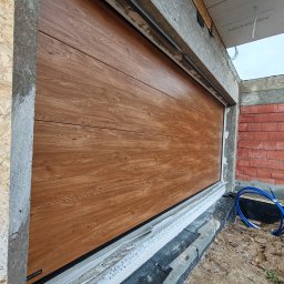 VINSTA SPÓŁKA CYWILNA WOJCIECH PAJĄK, DMYTRO ZHULINSKYI - Zamknięta brama garażowa segmentowa z panelami imitującymi drewno, widoczne detale montażu w surowym stanie, elementy izolacji i konstrukcji budynku.