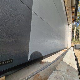 VINSTA SPÓŁKA CYWILNA WOJCIECH PAJĄK, DMYTRO ZHULINSKYI - Szara segmentowa brama garażowa z fakturą drewna, widoczne logo 'KRISHOME' w lewym dolnym rogu, z zewnątrz, na etapie montażu, z elementami konstrukcyjnymi w tle.