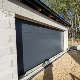 VINSTA SPÓŁKA CYWILNA WOJCIECH PAJĄK, DMYTRO ZHULINSKYI - Nowa, segmentowa brama garażowa w kolorze antracytowym, zamontowana w surowym budynku z białych bloczków, widoczne przygotowane materiały budowlane na zewnątrz.