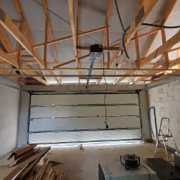 Wnętrze garażu w trakcie montażu segmentowej bramy garażowej, widoczny napęd elektryczny pod sufitem i konstrukcja drewniana dachu.