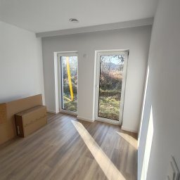 VINSTA SPÓŁKA CYWILNA WOJCIECH PAJĄK, DMYTRO ZHULINSKYI - Wnętrze nowo wykończonego pokoju z dwoma białymi oknami, przez jedno z nich widać żółtą rurę wentylacyjną, na podłodze panele w odcieniach beżu, przy ścianie kartony.