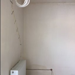 VINSTA SPÓŁKA CYWILNA WOJCIECH PAJĄK, DMYTRO ZHULINSKYI - Surowy stan wnętrza: biały grzejnik, wystające kable elektryczne w białej osłonie przymocowane do ściany, gładzie na ścianach w trakcie prac wykończeniowych, widoczne ślady po instalacji elektrycznej.