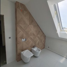 VINSTA SPÓŁKA CYWILNA WOJCIECH PAJĄK, DMYTRO ZHULINSKYI - Nowoczesna łazienka na poddaszu ze skośnym oknem dachowym, drewnianą ścianą z geometrycznym wzorem, białą ceramiką sanitarną (toaleta zakryta folią ochronną i bidet) oraz jasnymi, błyszczącymi...