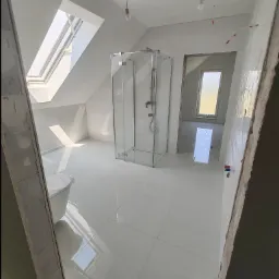 Łazienka na poddaszu w trakcie wykańczania: białe płytki podłogowe, kabina prysznicowa ze szkła, okno dachowe wpuszczające światło, surowy stan ścian z widocznymi instalacjami, widok na kolejne...