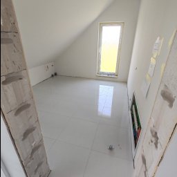 VINSTA SPÓŁKA CYWILNA WOJCIECH PAJĄK, DMYTRO ZHULINSKYI - Poddasze z białymi płytkami podłogowymi, oknem połaciowym zabezpieczonym folią i fragmentem instalacji elektrycznej w ścianie, widok przez surową ościeżnicę.