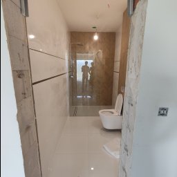 VINSTA SPÓŁKA CYWILNA WOJCIECH PAJĄK, DMYTRO ZHULINSKYI - Wnętrze łazienki w trakcie remontu, widoczne białe płytki, toaleta wisząca i kabina prysznicowa z drewnopodobną ścianą w jodełkę, surowy otwór drzwiowy z metalową ramą.