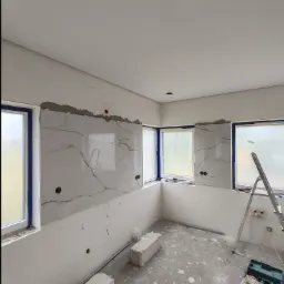 Montaż marmurowych płyt na ścianie w pomieszczeniu z trzema oknami, widoczne oklejone ramy okienne, drabina, resztki zaprawy i narzędzia na podłodze.