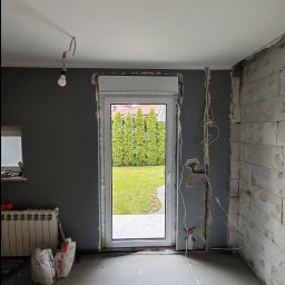 VINSTA SPÓŁKA CYWILNA WOJCIECH PAJĄK, DMYTRO ZHULINSKYI - Wnętrze w trakcie remontu z nowo zamontowanym oknem z roletą zewnętrzną, widoczne przewody elektryczne, surowa ściana z betonu komórkowego, grzejnik i worki z zaprawą.