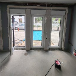 VINSTA SPÓŁKA CYWILNA WOJCIECH PAJĄK, DMYTRO ZHULINSKYI - Montaż okien: trzy białe okna z roletami zewnętrznymi w surowym stanie, jedno okno otwarte, widoczne przewody elektryczne i przedłużacz na podłodze.