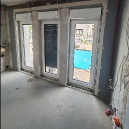 Wnętrze w trakcie remontu z trzema oknami z roletami zewnętrznymi, widoczne ślady prac wykończeniowych na ścianach i podłodze, instalacja elektryczna w trakcie montażu.